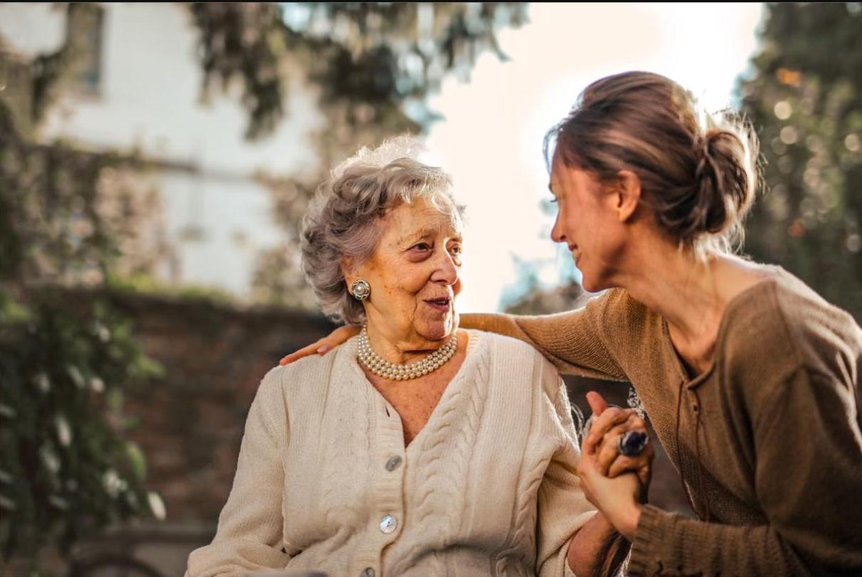 An adult daughter leaning in to talk with her mother in a sunlit garden